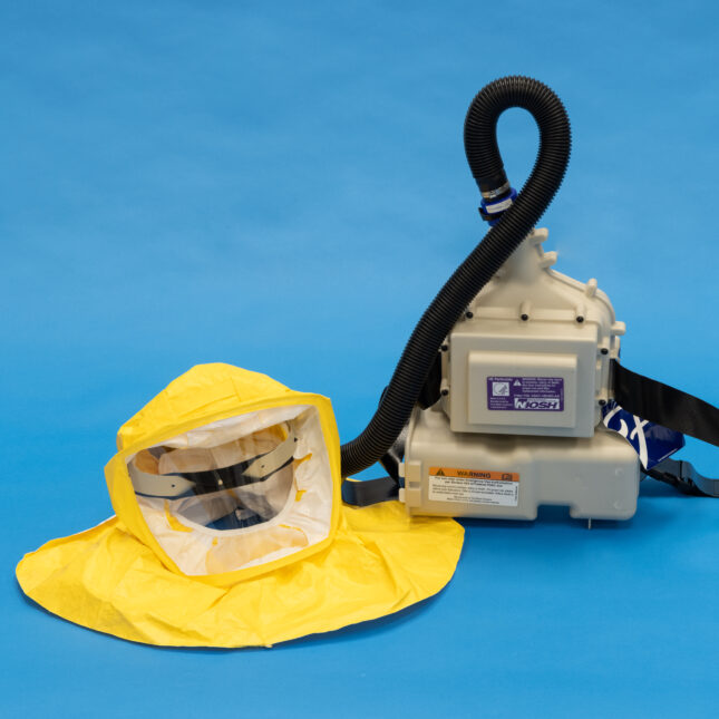 A powered air purifying respirator, including a helmet covered by a yellow hood on the left connected through a tube to a powered air system, placed on a blue floor — first opinion coverage from STAT