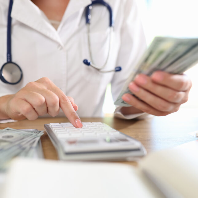 A doctor, with stethoscope around their neck, holds cash while operating a calculator — business coverage from STAT