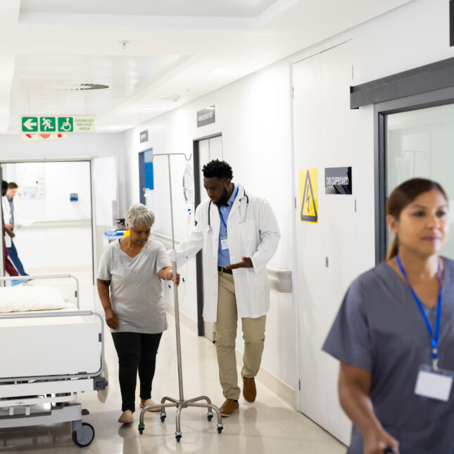 A doctor holds the arm of a senior patient with IV drip walking in corridor — hospitals coverage from STAT