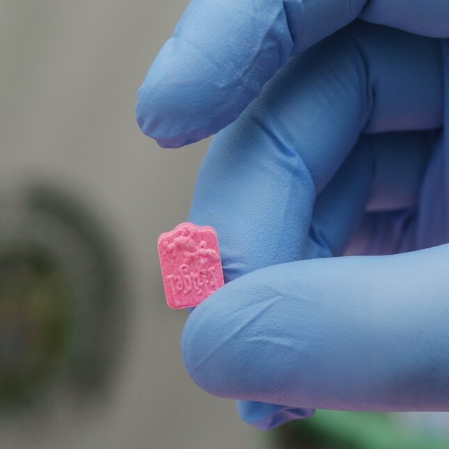 A hand in blue glove holds a pink MDMA tablet — health coverage from STAT