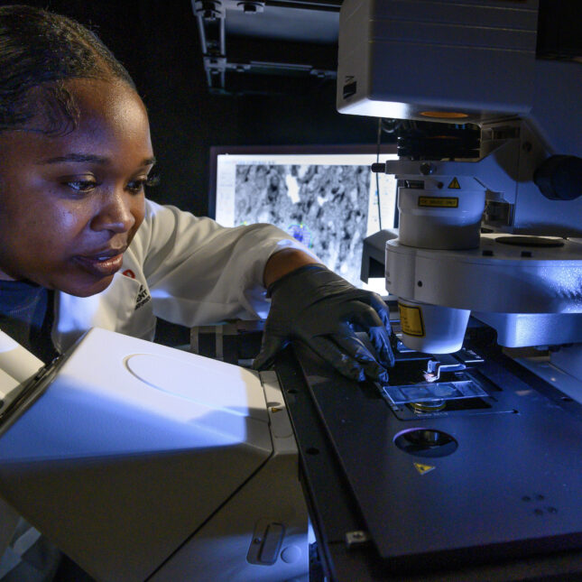 A Black person, wearing gloves, slides a piece of specimen under a microscope in a lab — coverage from STAT