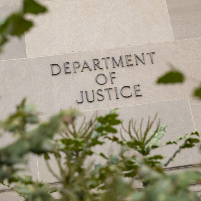 A sign that reads "Department of Justice" on its building behind some bushes — insurance coverage from STAT