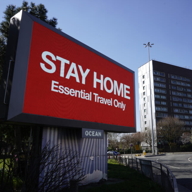 A digital board displays red background with white text that reads "STAY HOME" on the top and "Essential Travel Only" on the bottom — first opinion coverage from STAT