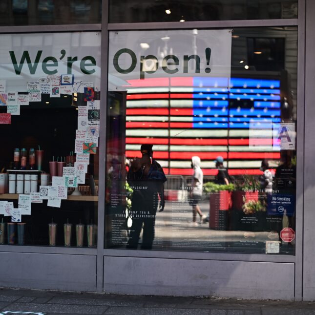 A glass window with coffee bottle display reflects the American flag and shows a banner that reads "We're Open!" — first opinion coverage from STAT