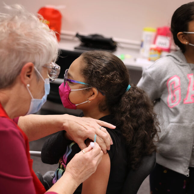 A child wearing glasses and a pink mask receives a vaccine in their left arm from an elderly medical staff member. Another masked child in a gray jacket stands in the background, holding a piece of green paper — first opinion coverage from STAT