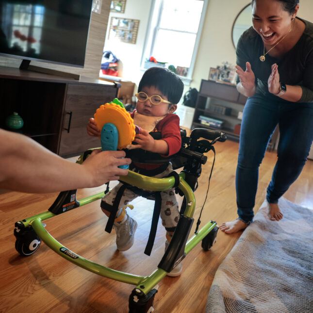 Nathan Guo, left, and his wife Ada Lio encourage their 17-month-old son, Lucas Guo, to take steps in his gate trainer at their home in Somerville, MA on February 16, 2024. Lucas has one of the world's rarest diseases, ZTTK Syndrome