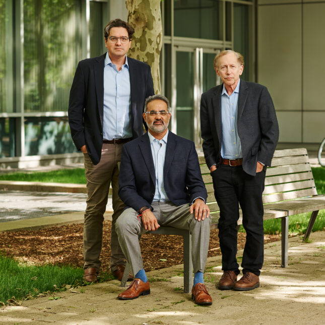 Three people in different suits, two standing up and the middle one sitting on a bench, looks to the camera — biotech coverage from STAT